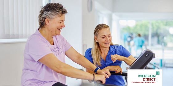 Eine Therapeutin zeigt einer Patientin, die auf einem Ergometer sitzt, wie sie verschiedene Einstellungen am Display verändern kann.