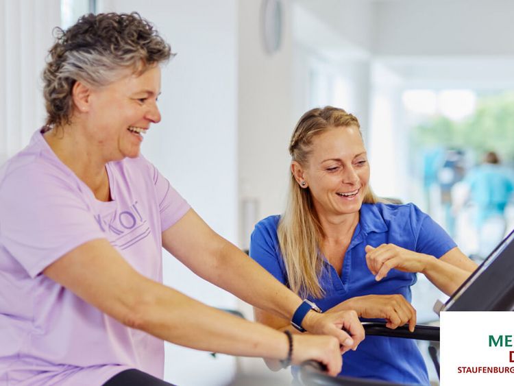 Eine Therapeutin zeigt einer Patientin, die auf einem Ergometer sitzt, wie sie verschiedene Einstellungen am Display verändern kann.