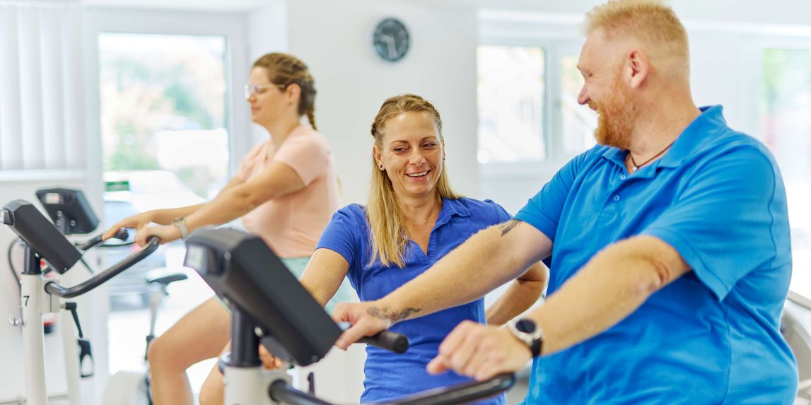 Im Sportpark der Klinik zeigt eine Therapeutin einem Patienten, wie er den Ergometer am besten nutzt